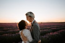 Vista lateral do casal recém-casado romântico em pé face a face no campo espaçoso contra o céu púrpura por do sol — Fotografia de Stock