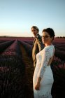 Braut und Bräutigam halten Händchen und gehen in blühendes Lavendelfeld und schauen in die Kamera — Stockfoto