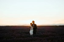 Vista lateral do casal recém-casado romântico em pé face a face no campo espaçoso contra o céu púrpura do pôr do sol olhando para a câmera — Fotografia de Stock