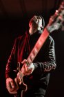 Низкий угол игры рок-гитариста на электрогитаре во время выступления в темной студии с красным светом — стоковое фото