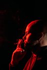 Vue latérale du rocker mâle avec tête chauve fumant et expirant en studio sombre avec lumière au néon rouge sur fond noir — Photo de stock