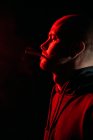 Side view of male rocker with bald head smoking and exhaling fume in dark studio with red neon light on black background — Stock Photo