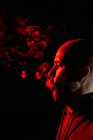 Vue latérale du rocker mâle avec tête chauve fumant et expirant en studio sombre avec lumière au néon rouge sur fond noir — Photo de stock