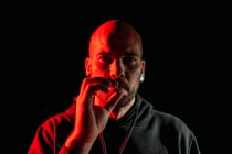 Sério roqueiro masculino fumar cigarro e olhando para a câmera no fundo preto em estúdio com iluminação vermelha — Fotografia de Stock