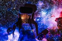 Menina jovem irreconhecível em desgaste casual e fone de ouvido VR recebendo nova experiência e tocando objeto virtual no quarto com iluminação de projetor colorido — Fotografia de Stock