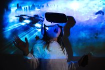 Unbekanntes junges Mädchen in Freizeitkleidung und VR-Headset macht neue Erfahrungen und berührt virtuelles Objekt im Raum mit farbenfroher Projektor-Beleuchtung — Stockfoto