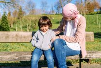 Нежная спокойная мать с раком в розовом шарфе на голове успокаивает грустную маленькую дочь, сидящую на скамейке в зеленом парке — стоковое фото