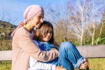 Счастливая мать с раком в розовом шарфе на голове, обнимающая молодую дочь, сидящую на скамейке в зеленом парке и отворачивающуюся — стоковое фото