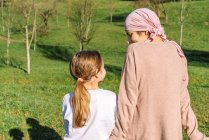 Vista posterior de la tierna madre tranquila con cáncer con pañuelo rosa en la cabeza hablando con la pequeña hija sentada en un banco en el parque verde - foto de stock