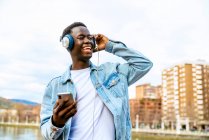 Young positive black male with cellphone listening to song from headphones while looking up on urban embankment — Stock Photo