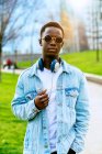 Contenido joven Hombre afroamericano en chaqueta de mezclilla con auriculares en el camino entre el césped en la ciudad - foto de stock