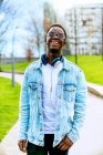 Jovem conteúdo afro-americano masculino em jaqueta de ganga com fones de ouvido no caminho entre gramados na cidade — Fotografia de Stock
