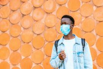 Hombre negro contemplativo joven irreconocible en chaqueta de mezclilla y máscara respiratoria mirando hacia otro lado durante la pandemia de coronavirus - foto de stock