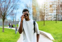 Young African American man wearing a t-shirt and sunglasses on a path between lawns in the city. — Stock Photo