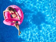 Зверху жінка в купальнику плаває на рожевому надувному кільці в прозорій блакитній воді відкритого басейну під час літніх канікул — стокове фото
