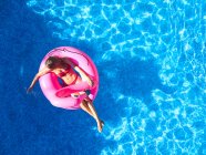 Зверху жінка в купальнику плаває на рожевому надувному кільці в прозорій блакитній воді відкритого басейну під час літніх канікул — стокове фото