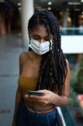 Portrait of attractive young afro latin woman wearing a facemask and using smartphone in a commercial mall, Colombia — Stock Photo