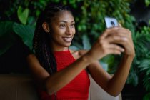 Ritratto di attraente giovane donna afro latino con dreadlocks in un top rosso all'uncinetto utilizzando smartphone in tavolo ristorante, Colombia — Foto stock