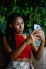 Portrait de attrayant jeune femme afro latine avec dreadlocks dans un crochet haut rouge en utilisant smartphone dans la table du restaurant, Colombie — Photo de stock