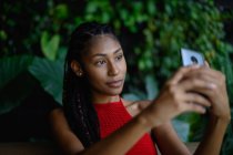 Portrait of attractive young afro latin woman with dreadlocks in a crochet red top using smartphone in restaurant table, Colombia — Stock Photo