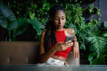 Portrait de attrayant jeune femme afro latine avec dreadlocks dans un crochet haut rouge en utilisant smartphone dans la table du restaurant, Colombie — Photo de stock