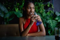 Attrayant jeune femme afro latine avec dreadlocks dans un crochet rouge top boit de l'eau au restaurant, Colombie — Photo de stock