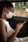 Jovem afro mulher latina na máscara facial usa smartphone no banco de trás de um carro — Fotografia de Stock