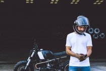 Full body of serious young ethnic male biker in casual clothes and cap standing near motorcycle near industrial cranes at seaside — Stock Photo