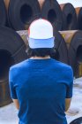 Вид ззаду на невпізнаваного чоловіка з темним волоссям у повсякденному вбранні та кепці, що стоїть біля стопки металевих листів котушок на промисловому заводі — стокове фото