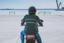 Rückansicht eines unkenntlich gemachten männlichen Bikers in lässigem Outfit und Schutzhelm, der auf einem Motorrad auf einer Böschung in Seenähe an einer Baustelle sitzt — Stockfoto