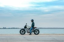 Вид сбоку на неузнаваемого байкера в повседневной одежде и защитном шлеме, сидящего на мотоцикле на набережной возле моря на строительной площадке — стоковое фото