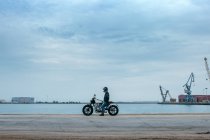 Vista laterale di biker maschio irriconoscibile in abbigliamento casual e casco protettivo seduto su moto sul terrapieno vicino al mare in cantiere — Foto stock