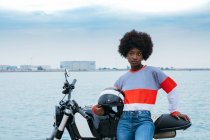 Concentrated young black female biker with Afro hair in trendy outfit and helmet while sitting on motorcycle at seaside — Stock Photo