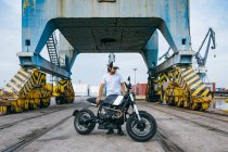 Full body of serious young ethnic male biker in casual clothes and cap standing near motorcycle near industrial cranes at seaside — Stock Photo