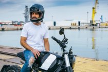 Уверенный молодой бородатый этнический парень в белой футболке и джинсах, стоящий на мотоцикле на берегу моря — стоковое фото