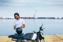 Confiant jeune homme ethnique barbu en t-shirt blanc et jeans en moto debout au bord de la mer — Photo de stock