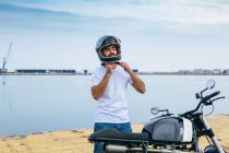 Confiado joven barbudo étnico en camiseta blanca y jeans atando casco mientras está de pie motocicleta en la playa - foto de stock