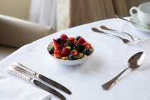 Сверху миска здорового мюсли, увенчанная свежими сортами ягод, подаваемых на белом столе с серебром и чашкой кофе во время завтрака — стоковое фото
