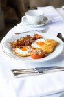 Высокий угол аппетитных яиц с ломтиками бекона подается на тарелке с фаршированным помидором и хрустящей моцареллой и помещается на стол с катлером и чашкой кофе во время завтрака — стоковое фото