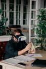Chef masculin méconnaissable en uniforme assis à table avec divers documents et smartphone et tenant du maïs grillé tout en faisant l'expérience de la réalité virtuelle dans un casque VR au restaurant — Photo de stock