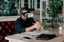 Unerkennbarer männlicher Koch in Uniform sitzt mit diversen Dokumenten und Smartphone am Tisch und hält gegrilltes Mais in der Hand, während er Virtual Reality im VR-Headset im Restaurant erlebt — Stockfoto