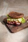 Alto ángulo de bocadillo apetitoso con pan crujiente fresco sobre hojas de lechuga y tocino sobre tabla de madera - foto de stock