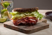 Високий кут апетитного бутерброда зі свіжим грубим хлібом над листям салату та беконом на дерев'яній дошці — стокове фото