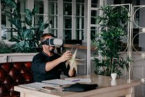 Неузнаваемый шеф-повар в форме сидит за столом с различными документами и смартфоном и держит жареную кукурузу, испытывая виртуальную реальность в виртуальной реальности в гарнитуре VR в ресторане — стоковое фото