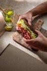 Desde arriba de cultivo cliente anónimo de restaurante comiendo sabroso sándwich hecho con tostadas de tocino y hojas de lechuga con agua con lima - foto de stock