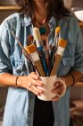 Cropped unrecognizable female master in casual clothes smiling and looking at camera while standing near shelf in workshop with mug of various paintbrushes in hand — Stock Photo