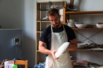 Concentrated young bearded male potter in casual clothes and apron creating white ceramic plate while working in studio — Stock Photo