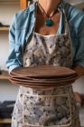 Ernte unkenntliche Kunsthandwerkerin in lässigem Outfit und Schürze hält Stapel flacher Tonteller während der Arbeit im Kunstatelier — Stockfoto