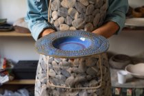 Crop unrecognizable artisan in apron demonstrating creative ceramic plate in workshop — Stock Photo