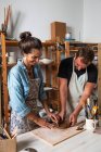 Positif femme d'âge moyen et jeune homme en vêtements décontractés et tabliers souriant tout en créant plaque d'argile pendant le cours de poterie en atelier — Photo de stock